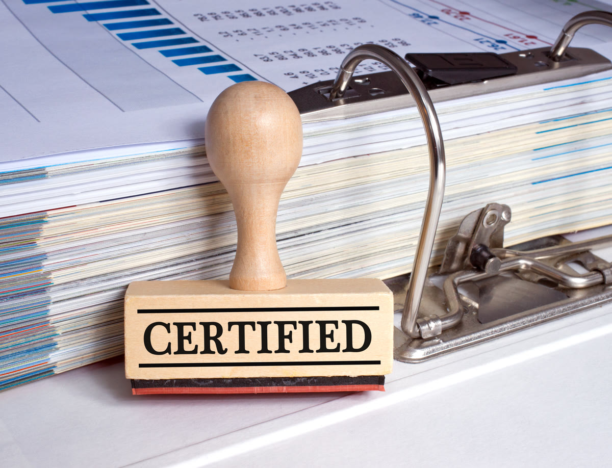 기업 서류가 편철된 바인더 옆에 CERTIFIED 문구가 새겨진 스탬프가 놓여 있는 모습입니다.